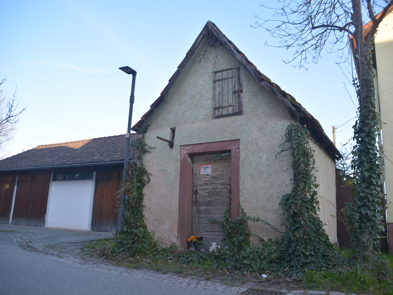 Wenn man vom Dorfplatz kommt wirkt das steinere Häuschen mit seiner Holztür und dem hölzernen Laden unterm Dach sehr verschlossen.