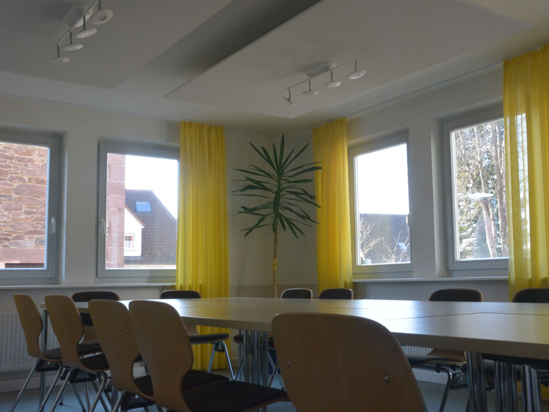 Große Fenster lassen Licht auf Tische und Stühle im Besprechungsraum in der Ortsverwaltung Neuhengstett fallen. Glebe Vorhänge säumen die Fenster; in der Ecke steht eine Palme.